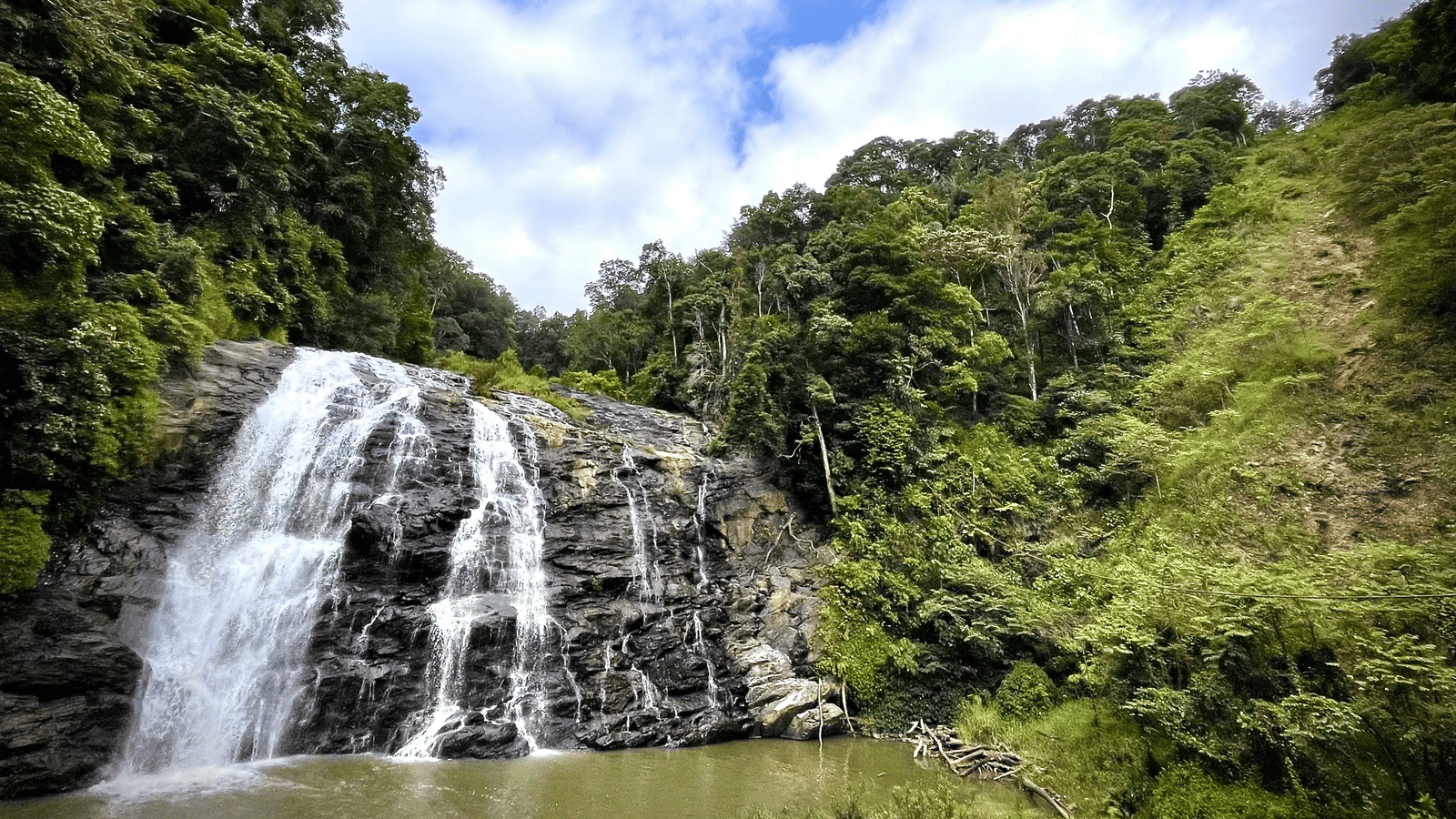 A waterfall flowing over a rocky cliff surrounded by trees and green vegetation under a partly cloudy sky.