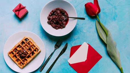A breakfast setting on a blue surface with a waffle, a bowl of cherries, a red envelope, a small red box, and a red tulip.