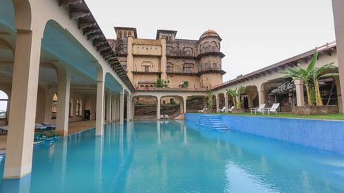 The swimming pool at Tijara Fort-Palace in Alwar, Rajasthan, reflects the historic architecture under an overcast sky.