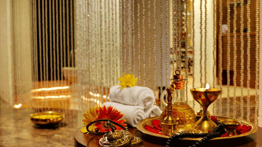 Myra Spa at Kenilworth Resort & Spa, Goa, featuring rolled white towels, a brass tray with massage oil, a lit lamp, fresh flowers on a round wooden table, and shimmering beaded curtains in the background.