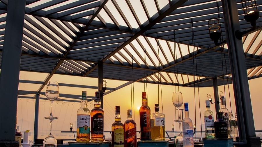  A close-up of a bar counter at sunset, with bottles of liquor backlit by the sun, and an outdoor rooftop setting.