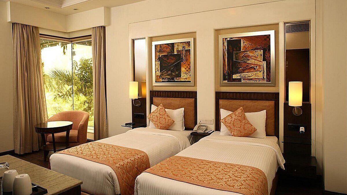 The Premium Twin Room offers twin beds with neatly arranged pillows, wooden bedside tables with lamps, a cushioned seating area with a wooden table, and large side windows for natural light at Inde Signature Tower, Gurgaon