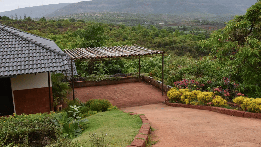 The surrounding landscape is lush and green, with trees, shrubs, and flowering plants. A paved pathway or driveway is visible, leading towards the covered structure.
