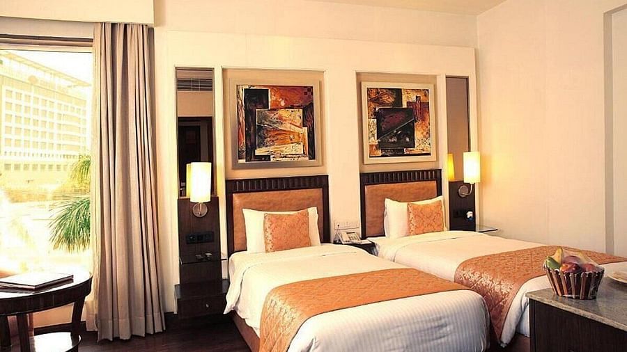 The Premium Twin Room offers twin beds with neatly arranged pillows, wooden bedside tables with lamps, a cushioned seating area with a wooden table, and large side windows for natural light at Inde Signature Tower, Gurgaon