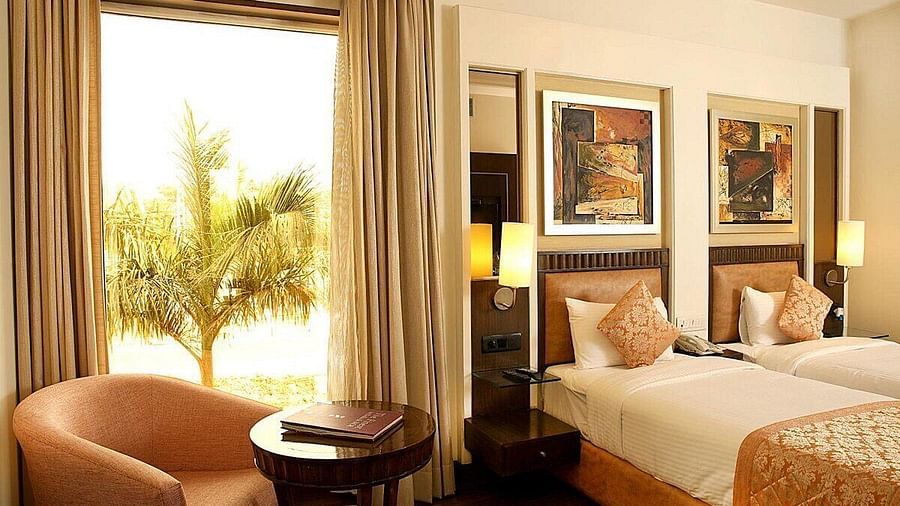 The Premium Twin Room offers twin beds with neatly arranged pillows, wooden bedside tables with lamps, a cushioned seating area with a wooden table, and large side windows for natural light at Inde Signature Tower, Gurgaon