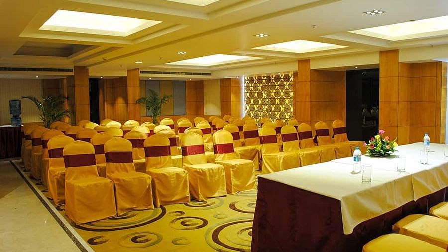 The image shows a conference room with rows of chairs facing a table with water bottles and flowers - Lords Eco Inn Navsari.