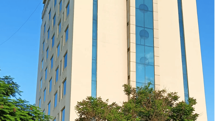 A tall building with multiple windows rises against a clear sky, partially framed by trees at its base. 