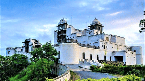 Majestic white fort perched on a hill, surrounded by lush greenery, under a partly cloudy blue sky.