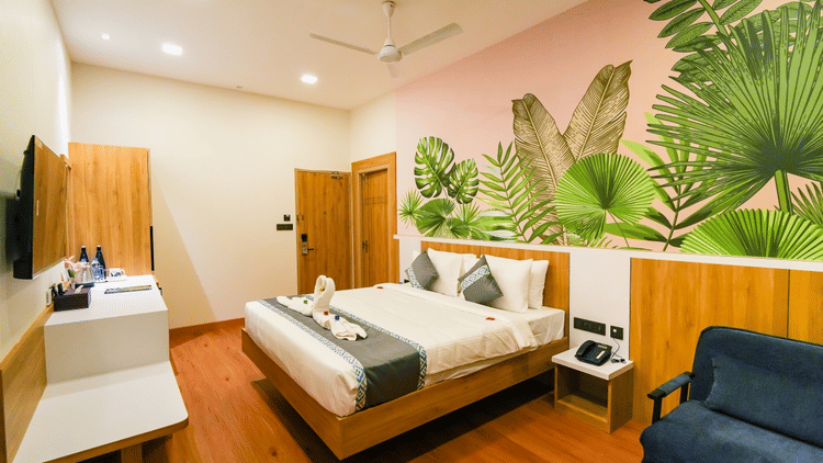 Modern hotel room with a double bed, tropical-themed wall art, wooden furnishings, a ceiling fan, and a comfortable seating area - MGM Vailankanni Residency.