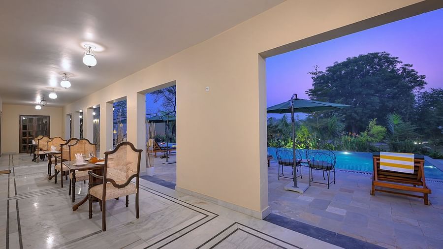 An open dining area with marble flooring and wooden chairs overlooks a poolside patio. Large openings frame the view of lush greenery, outdoor seating, and a twilight sky - Singhvilas, Ranthambore