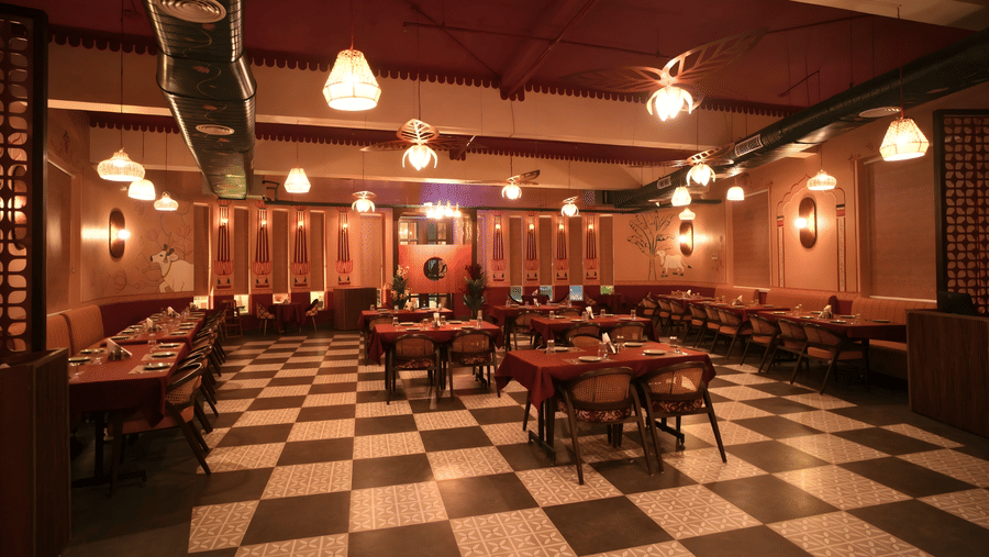 Side view of a warm, dimly lit restaurant interior featuring several tables covered in red tablecloths and a black and white checkered floor at Kadamb Kuteer, Vrindavan.
