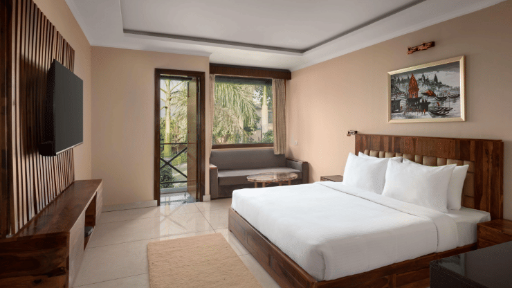 A Bay Room at Corbett Adventure Resort, Ramnagar, featuring a spacious bedroom with a king size bed, comfortable seating area, television, and large windows opening to lush outdoor views.