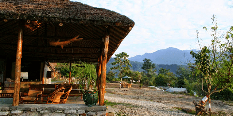 image shows a thatched-roof tent stay at the jamoon corbett, with a view of the mountains in the background.