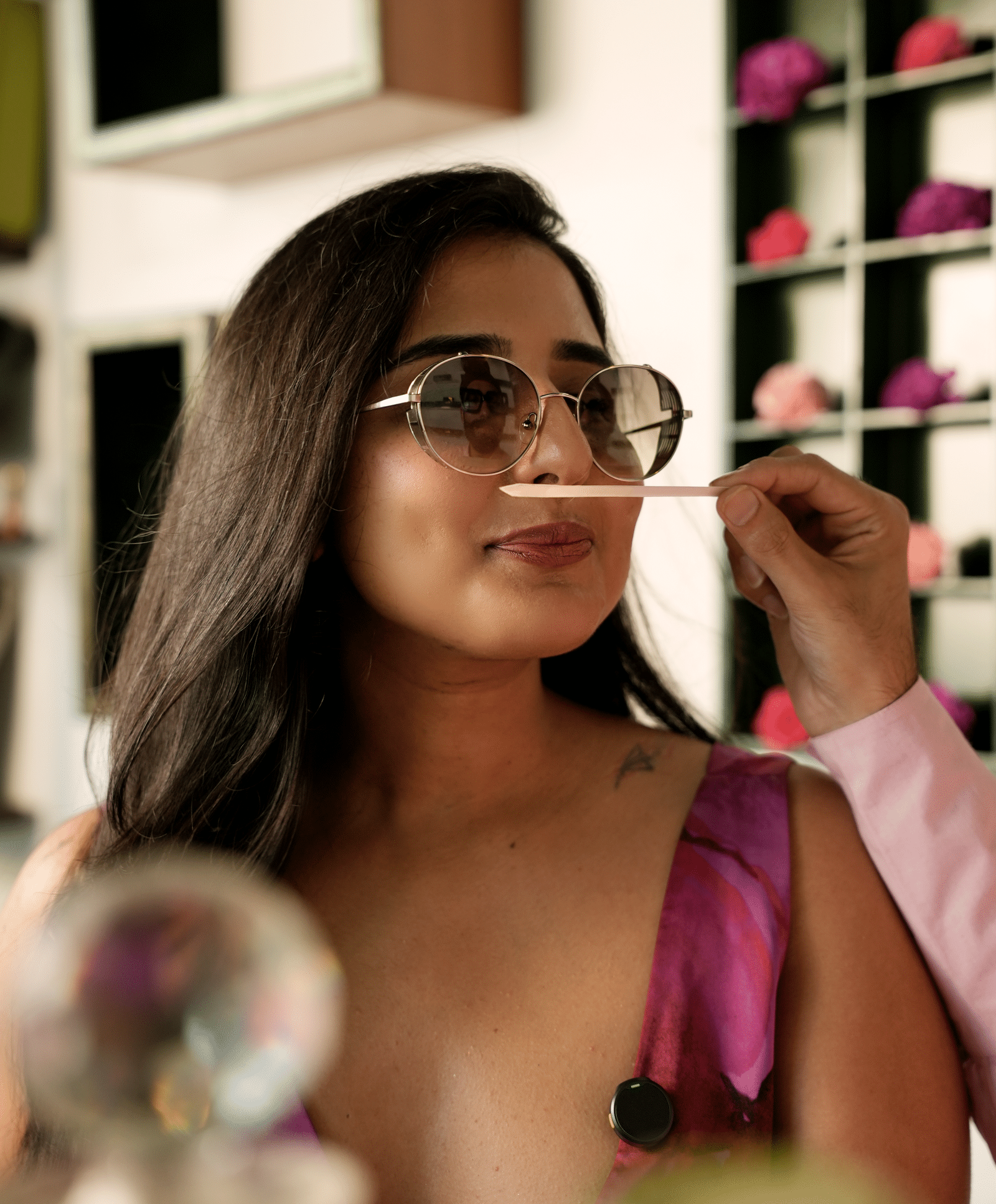 A woman wearing sunglasses and a vibrant pink dress smells a fragrance strip being held up to her nose by another person. She is in a stylish, modern setting with a display of colourful flowers in the background - Kastoor x Sawai Hospitality