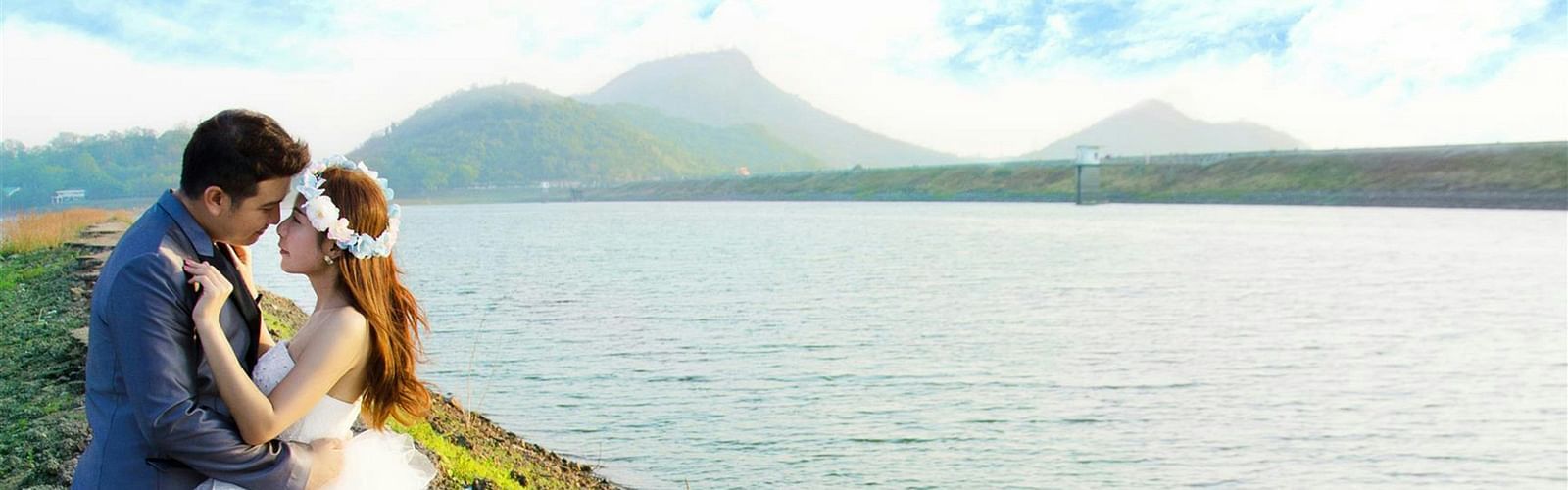 a bride and groom posing for a picture next to a waterbody with a mountain in the background