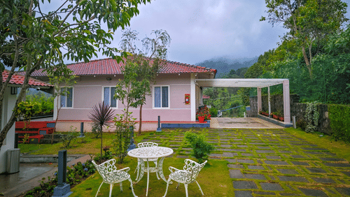 A modern white house with a red roof and a large carport, surrounded by a green lawn with a stone path and a white outdoor dining set, all under a cloudy sky with hills in the background.