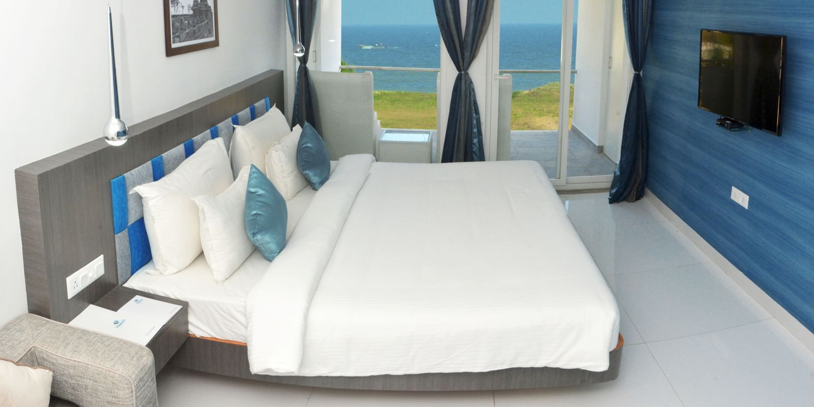 A hotel bedroom with a large white bed, blue accent wall, and a view of the outdoors through a large window | Grande Chalet Sea View | Grande Bay Resort & Spa, Mamallapuram