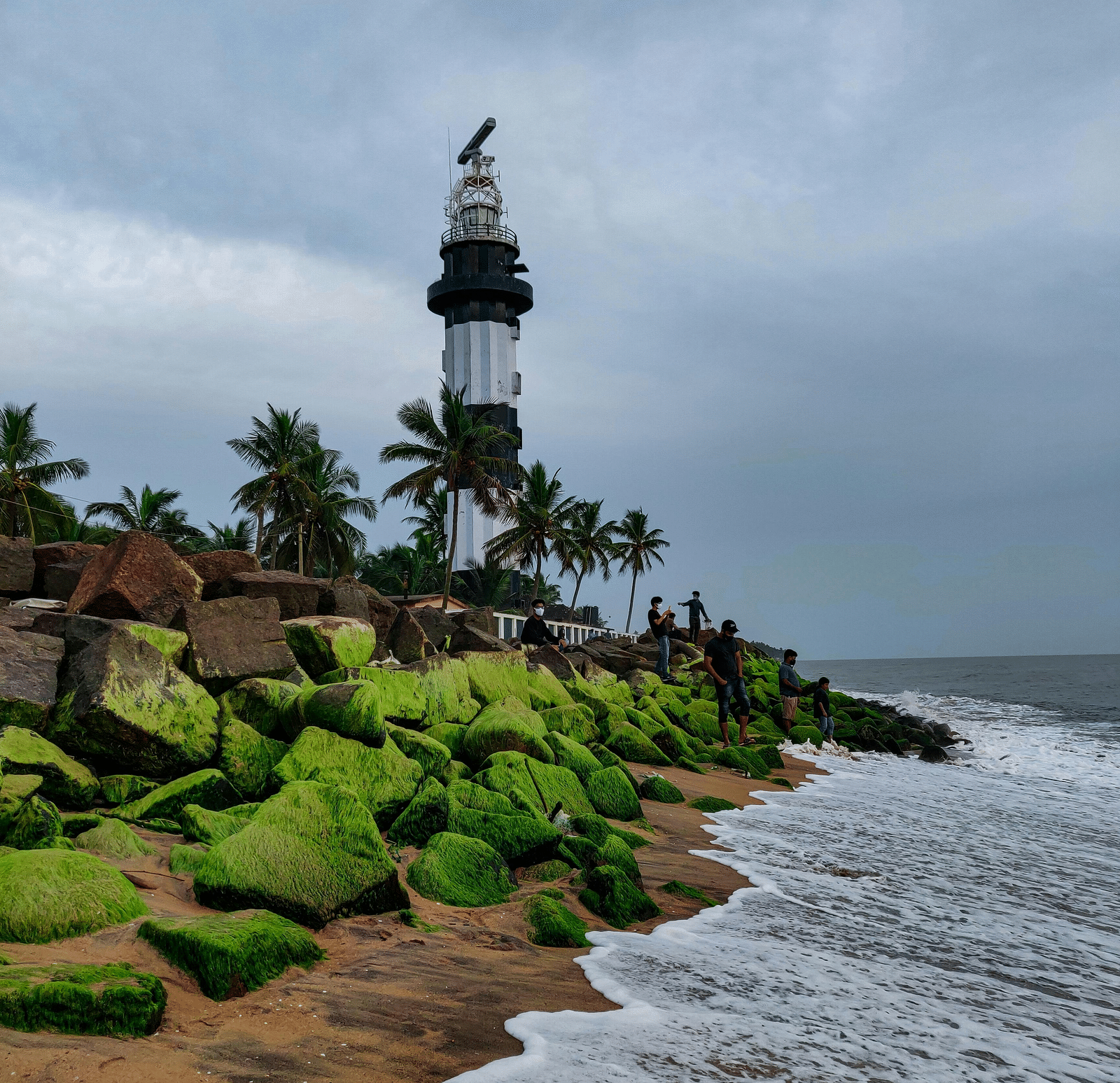 A scenic view of the Pondicherry Lighthouse standing tall amidst a rocky, moss-covered shoreline meeting the ocean under a cloudy sky. - Adventure things to do in Pondicherry