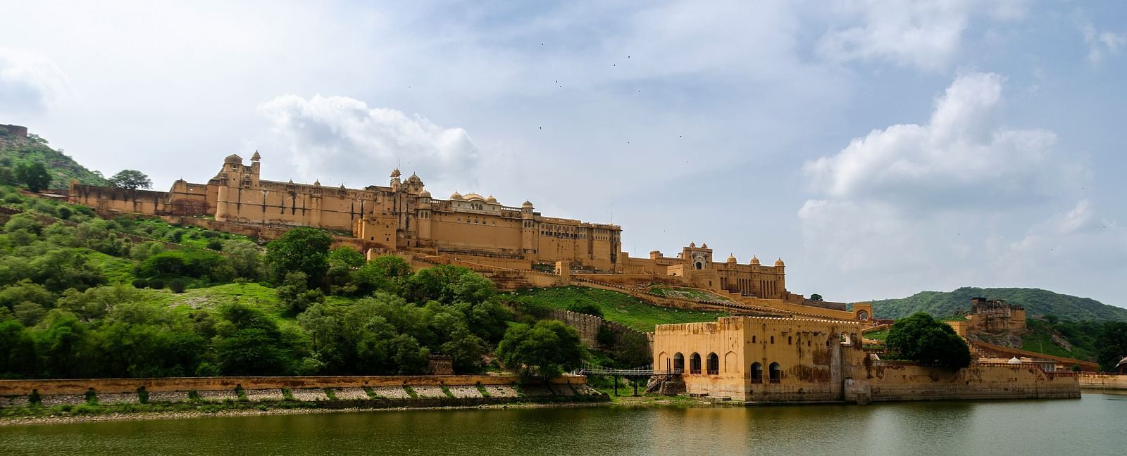 An imposing amber fort sprawls across a green hill, overlooking a serene lake under a cloudy sky.