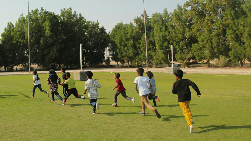 A group of children running around the large, open green field under a clear sky.