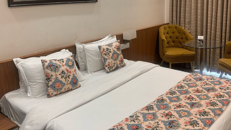 A Superior Room at the Prince ZENQ Xpress Resortt Rohtak, featuring a large bed with a floral runner, decorative pillows, a painting on the wall, and two velvet armchairs with a glass side table.