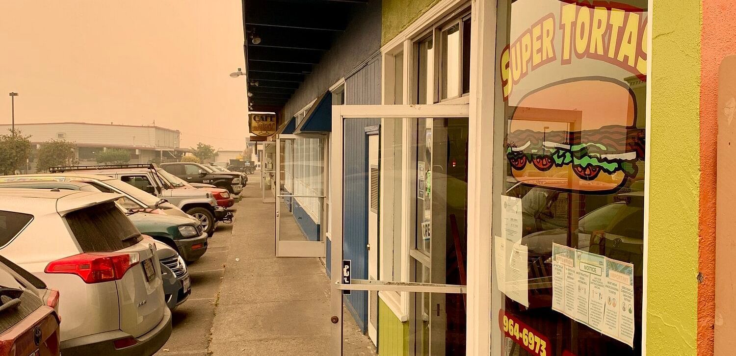 A Super Tortas restaurant with a bright green exterior features a hanging sign and window decals with a cartoon-style sandwich graphic. A row of parked cars lines the side of the building.