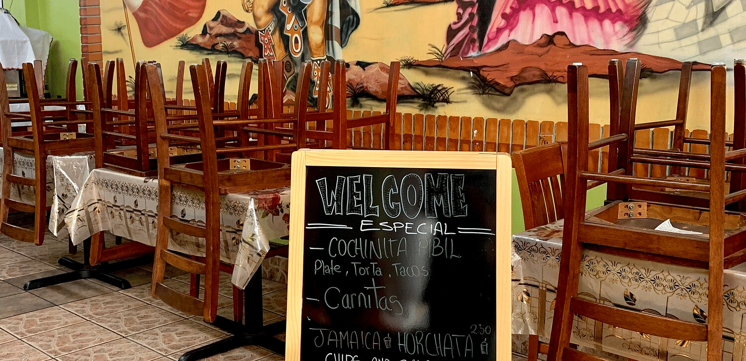 A chalkboard sign displaying specials like Cochinita Pibil and Carnitas sits in front of a restaurant interior with a large mural and empty tables.
