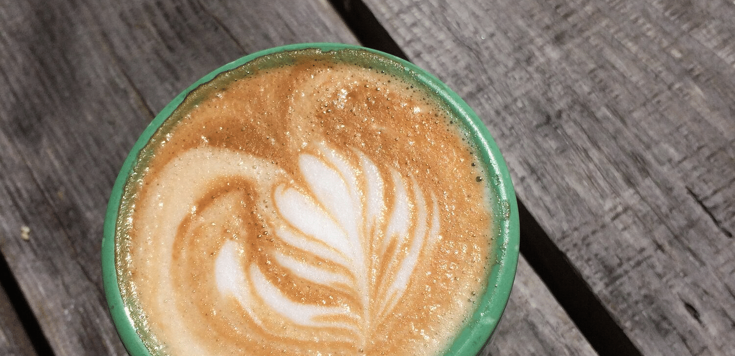 A green mug sits on a weathered wooden table, filled with a latte featuring a delicate leaf design in the foam.