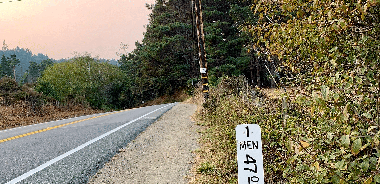 A roadside sign indicates 1 Man and 4700 ft against a backdrop of a telephone pole, road, and trees.