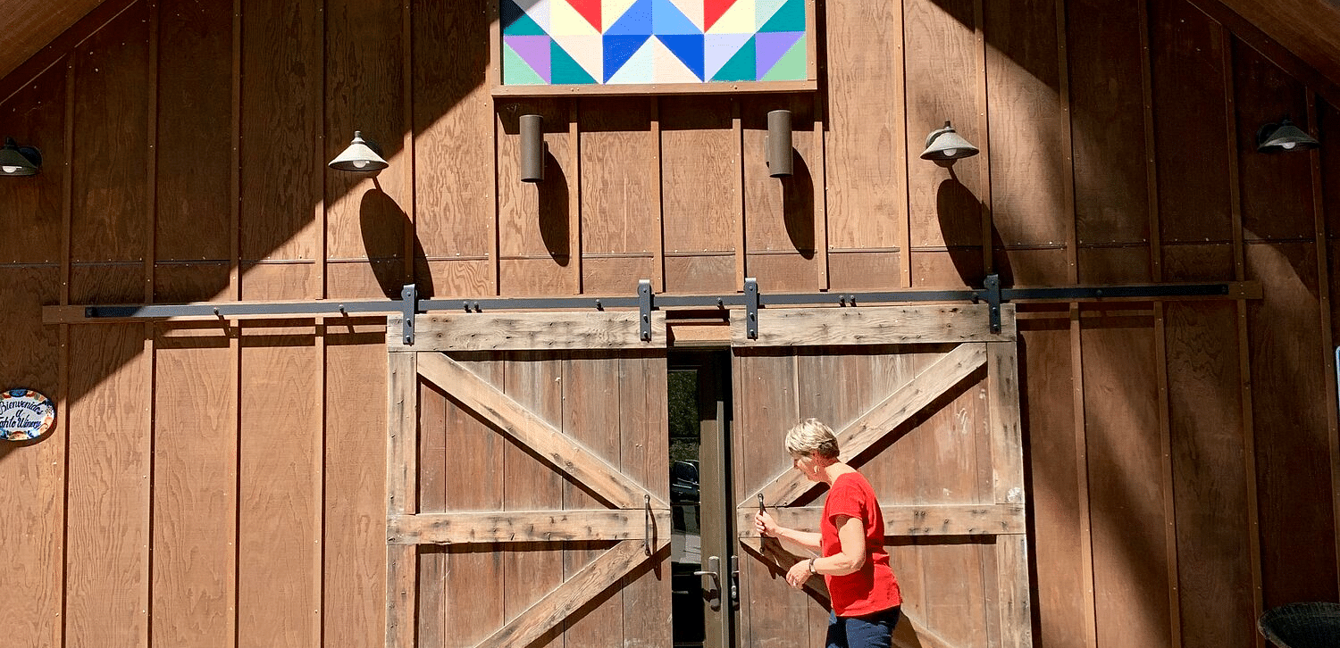  A building with a gabled roof and large barn doors, featuring a decorative quilt square above, with a person opening the doors.