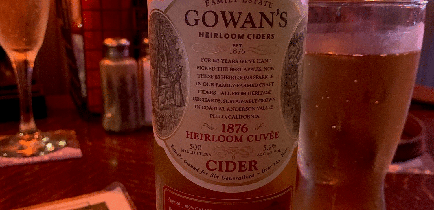 A bottle of Gowans cider stands on a table next to a glass of cider, with a menu and other items blurred in the background - Brewery Gulch Inn & Spa.