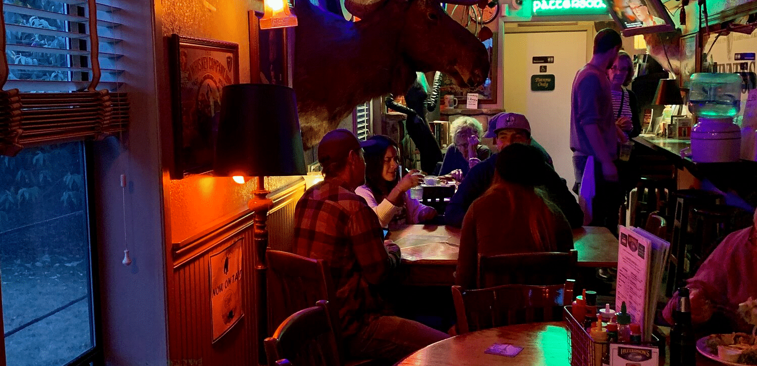 A dimly lit bar interior with patrons seated at tables, a Bass sign visible, and colorful lighting fixtures.