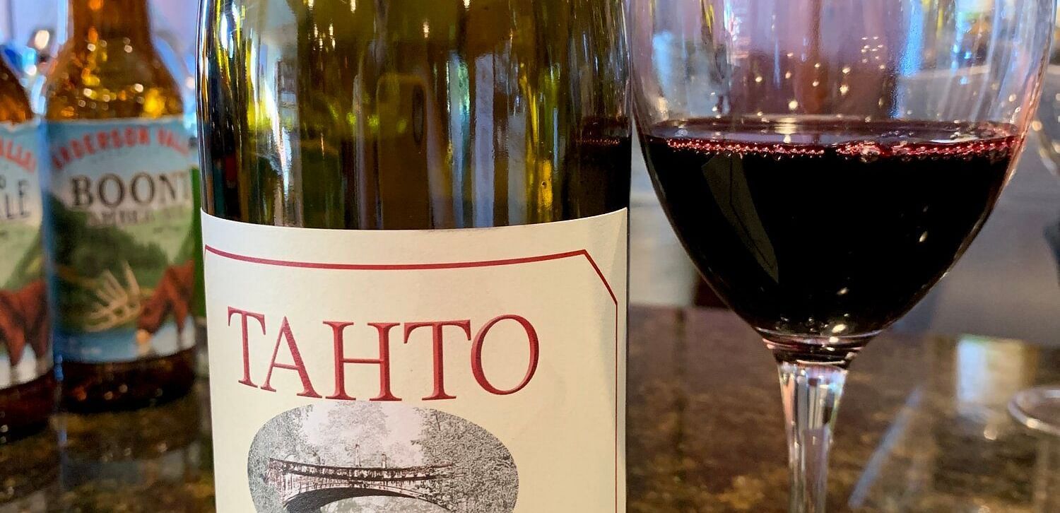  A bottle of Tahto Petite Sirah wine stands next to a stemmed glass filled with red wine, on a countertop.