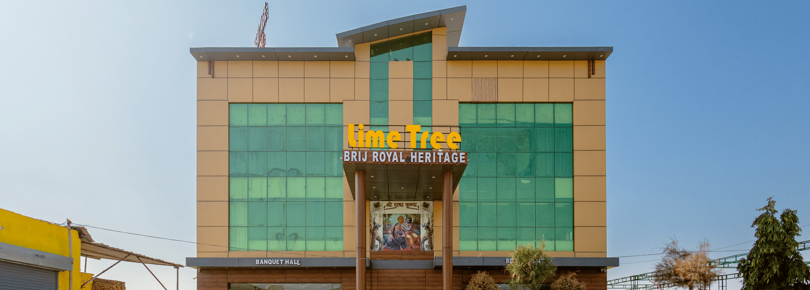 A daytime view of Lime Tree Hotels & Banquet, Vrindavan building with glass panels, yellow accents, and an open parking area in front.