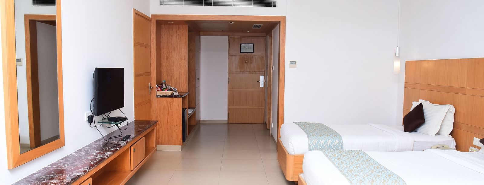 A well-furnished hotel room with twin beds, wooden interiors, and a sleek TV unit for added convenience.- Dhanalakshmi Srinivasan Hotels, Perambalur