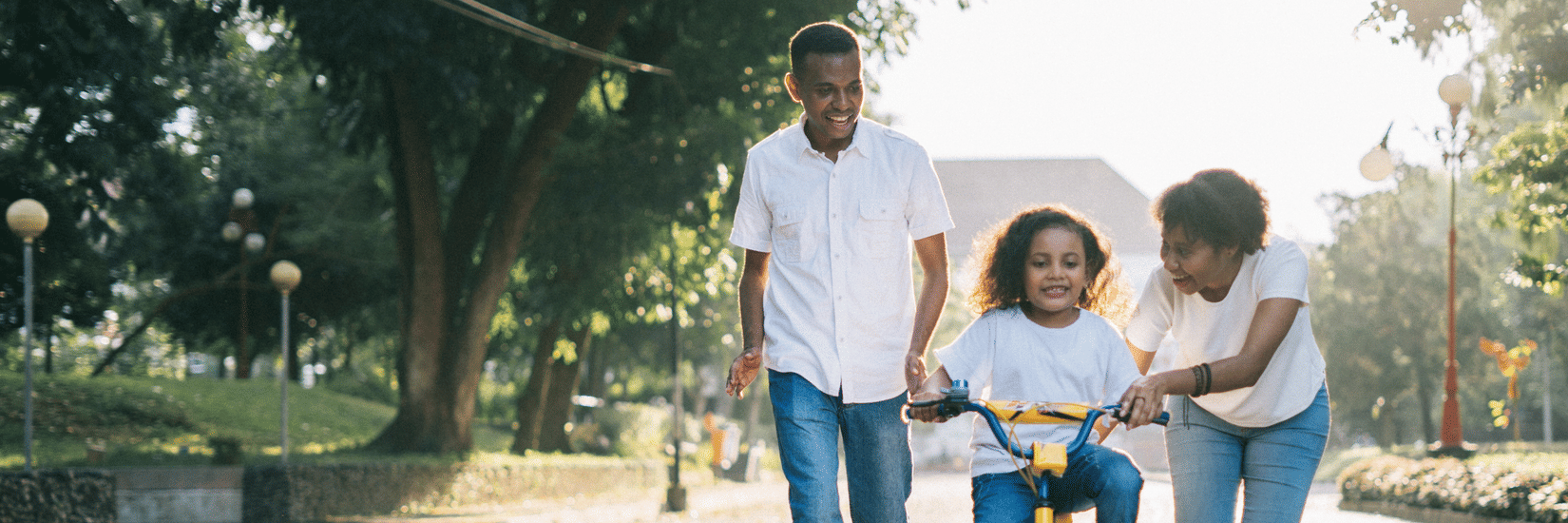 A mother and father smiling as they help their young daughter learn to ride a bicycle with training wheels on a sunny park path.