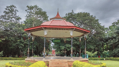 An open pavilion with a red roof surrounded by greenery and a walkway leading to a garden.