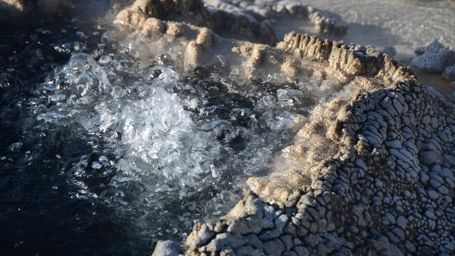 A close-up shot of boiling water bubbling vigorously within a geothermal pool bordered by rough, grey mineral deposits.