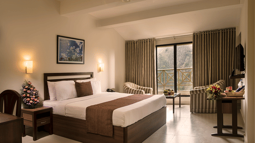 Room interior with a made-up bed featuring white linens and brown accents. A seating area with a sofa and a table is visible - Abad Copper Castle, Munnar