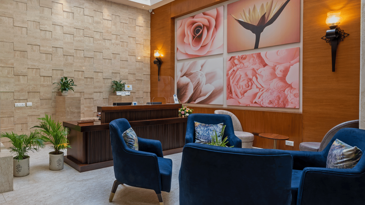 Hotel Saket 27 lounge with blue armchairs, wooden desk, potted plants, and large floral artwork on the wall.