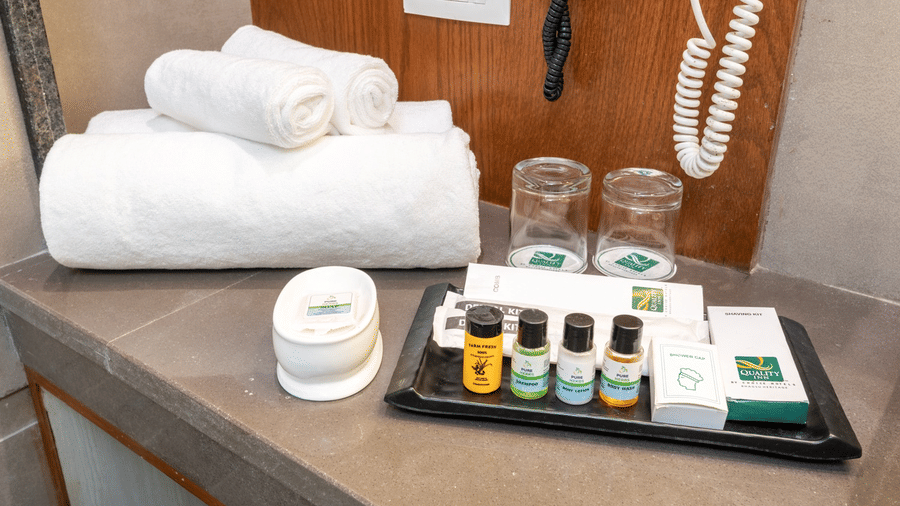 Bathroom amenity tray at Quality Inn Dharamshala with neatly folded white towels, glass tumblers, soap dish, and green labelled toiletry bottles on a grey counter.