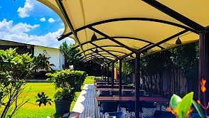 Covered outdoor dining area with tables lined under a beige canopy amidst lush greenery. | Vistar Resort, Bengaluru
