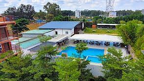 Aerial view of a resort complex featuring a central swimming pool surrounded by lush trees and buildings. | Vistar Resort, Bengaluru
