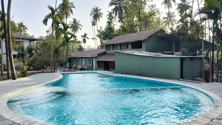 An outdoor swimming pool sits in front of a building surrounded by tall palm trees at Oceanature Resort, Alibaug.