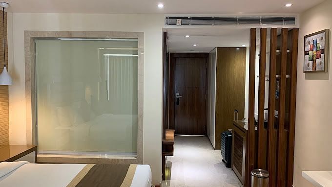 A hotel suite, featuring a sleek glass partition separating the bathroom area from the rest of the room, complemented by modern wooden accents and soft lighting at Yashshree The Imperial Inn, Siliguri.