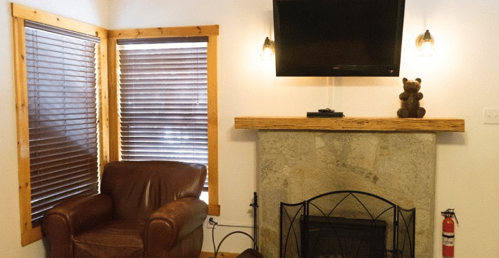 Overview of a Living area at Shaver Lake Village Hotel with a brown leather armchair, fireplace, TV, and blinds on two windows.