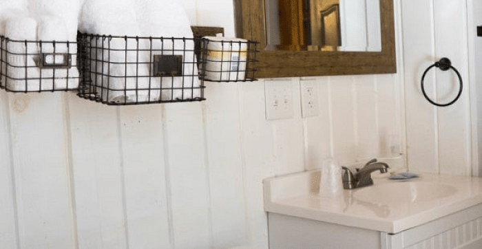 Overview of a Bathroom at Shaver Lake Village Hotel with white paneling, wire baskets holding towels, a wooden framed mirror, and a white vanity sink.