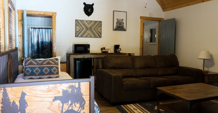 Overview of a Room at Shaver Lake Village Hotel with a brown couch, bed with decorative headboard, mini fridge, microwave, bear head decoration, and wood ceiling.
