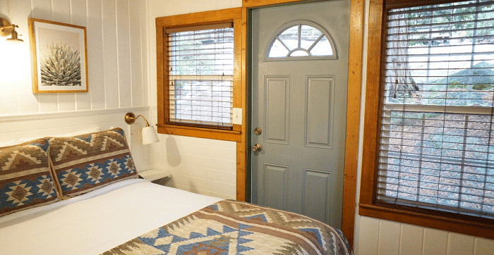 Overview of a Room at Shaver Lake Village Hotel with a bed featuring a patterned blanket, a grey door with a half-circle window, and two windows with blinds.