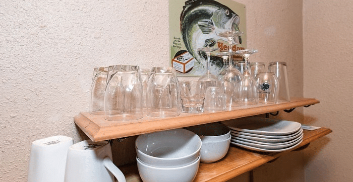 Overview of a kitchenette with a Shelf at Shaver Lake Village Hotel with glasses, bowls, plates, and mugs, below a painting of a fish.